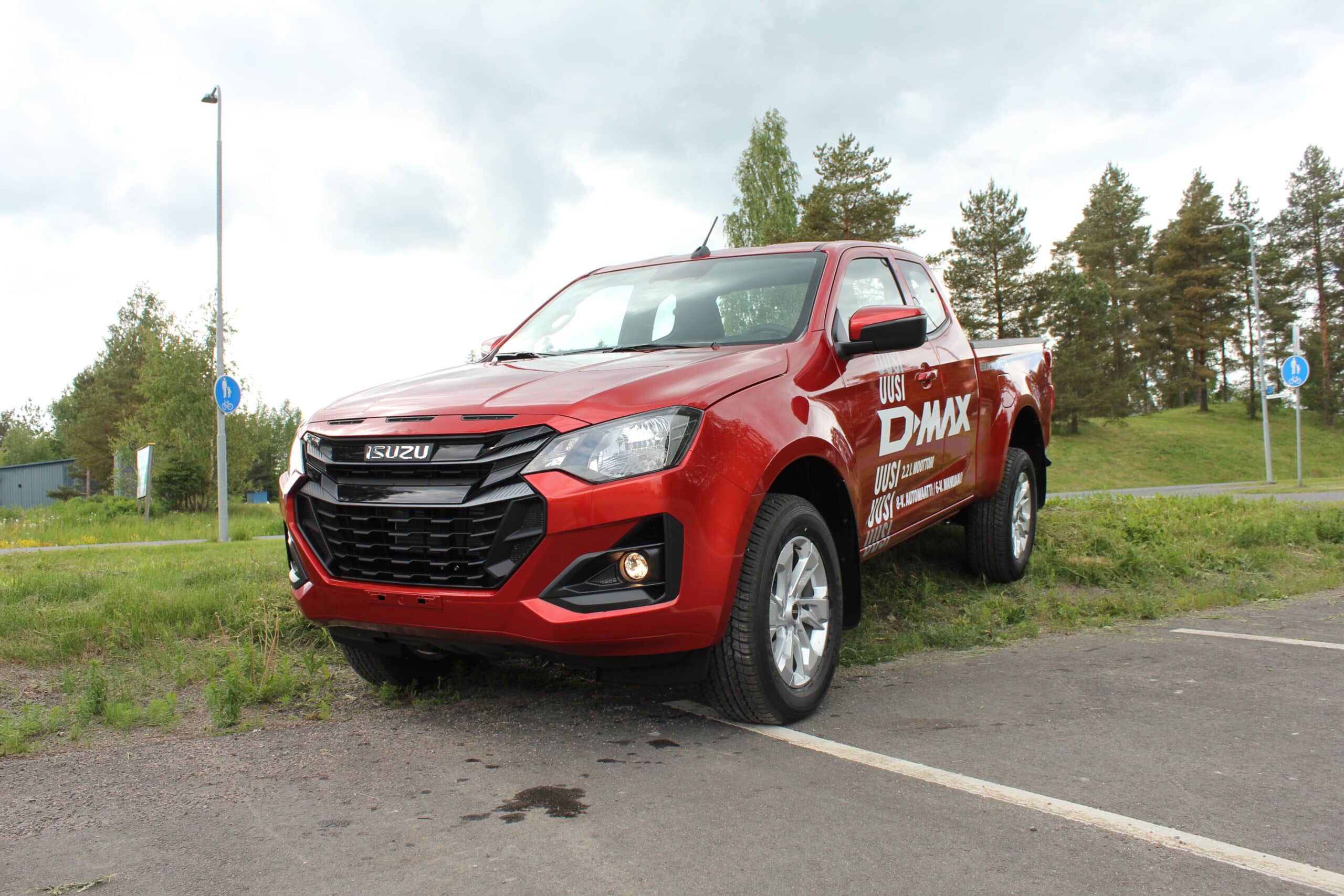 Red Isuzu D-Max pickup truck parked on a paved area with grass and trees in the background.